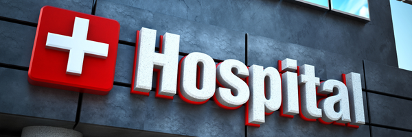 Medicare Part A: Modern hospital building sign with 3D white letters and a red cross emblem on a dark grey facade, signifying a healthcare facility.