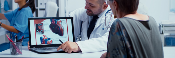 Medicare Part B: Male doctor in a white coat showing an elderly female patient a detailed human heart anatomy diagram on a laptop during a medical consultation about cardiac health.